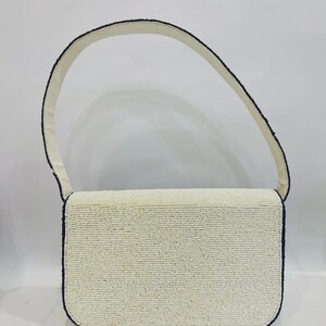 K&ouml;nnte beinhalten: Eine rechteckige, cremefarbene Handtasche mit passendem Trageriemen. Die Kanten sind mit schwarzen Perlen umrandet. Der Riemen ist cremefarben. Die Tasche ist ein klassisches, elegantes Accessoire.