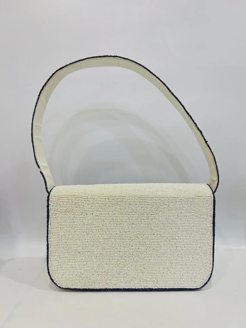 K&ouml;nnte beinhalten: Eine rechteckige, cremefarbene Handtasche mit passendem Trageriemen. Die Kanten sind mit schwarzen Perlen umrandet. Der Riemen ist cremefarben. Die Tasche ist ein klassisches, elegantes Accessoire.