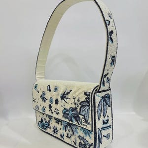 K&ouml;nnte beinhalten: Elfenbeinfarbene Handtasche mit Perlenbesatz und floralem Design in Blau- und Schwarzt&ouml;nen. Die Tasche hat einen Klappverschluss und einen passenden Perlenhenkel. Die Kanten sind schwarz umrandet. Es ist eine Schultertasche.