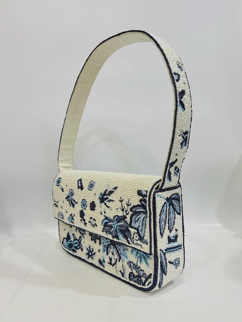 K&ouml;nnte beinhalten: Elfenbeinfarbene Handtasche mit Perlenbesatz und floralem Design in Blau- und Schwarzt&ouml;nen. Die Tasche hat einen Klappverschluss und einen passenden Perlenhenkel. Die Kanten sind schwarz umrandet. Es ist eine Schultertasche.