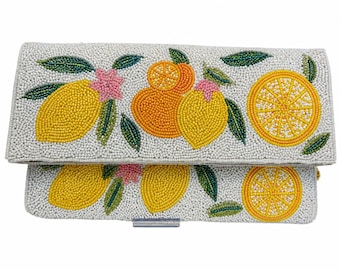 May include: A white beaded clutch featuring a colorful citrus design. The clutch is adorned with yellow lemons, orange oranges, pink flowers, and green leaves. The beaded pattern creates a textured surface, perfect for a summer accessory.