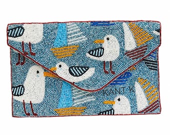 May include: A beaded clutch with a nautical theme. The clutch features a light blue background with white seagulls and sailboats in various colors. The edges are trimmed with a red border.