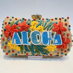 Op de afbeelding: Een rechthoekige clutch met het woord "ALOHA" in blauwe kralenletters. De tas is gemaakt van een beige geweven materiaal en versierd met rode, gele en groene bloemenborduursels. Een gouden sluiting sluit de bovenkant af.