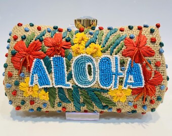 Puede incluir: Bolso de mano rectangular con la palabra "ALOHA" en letras de cuentas azules. El bolso está hecho de un material tejido de color beige y decorado con bordados florales rojos, amarillos y verdes. Un cierre dorado asegura la parte superior.
