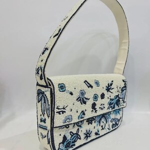 K&ouml;nnte beinhalten: Eine perlenbesetzte Handtasche mit cremefarbenem Grund, verziert mit blauen Blumen- und Bl&auml;ttermustern. Die Tasche hat einen Klappverschluss und einen passenden cremefarbenen Henkel. Die Kanten sind dunkelblau umrandet. Das Wort "KANTIK" ist unten rechts sichtbar.