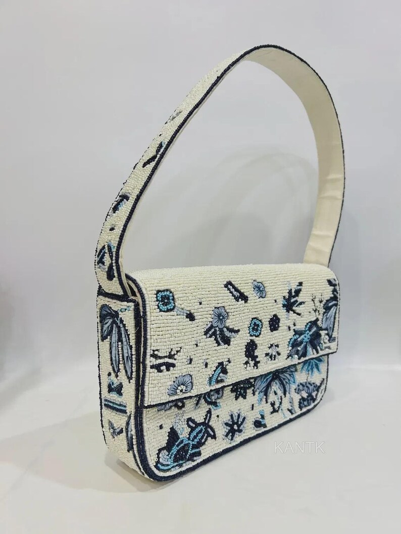 K&ouml;nnte beinhalten: Eine perlenbesetzte Handtasche mit cremefarbenem Grund, verziert mit blauen Blumen- und Bl&auml;ttermustern. Die Tasche hat einen Klappverschluss und einen passenden cremefarbenen Henkel. Die Kanten sind dunkelblau umrandet. Das Wort "KANTIK" ist unten rechts sichtbar.