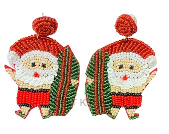 May include: A pair of Christmas-themed earrings featuring Santa Claus with a surfboard. The earrings are made of red, white, green, and beige beads. The Santa Claus figures have red hats and shorts, white beards, and hold green surfboards.