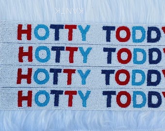 May include: Four beaded lanyards with the words "HOTTY TODDY" in red, blue, and white beads. Each lanyard has a silver-colored clasp. The lanyards are displayed on a white, fluffy surface.