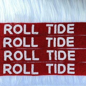 May include: Four red beaded straps with the words "ROLL TIDE" in white beads. Each strap has a white star at the end and silver-colored clasps. The straps are displayed on a white, textured surface.