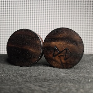 May include: Two dark brown wooden ear plugs. One plug has a carved symbol. The plugs are round and have a visible wood grain pattern. The background is a light gray and white checkered pattern.