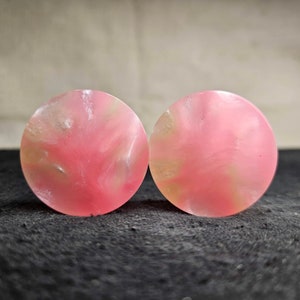 May include: Two round, pink ear plugs with a marbled effect. The plugs have a swirl of lighter pink and yellow tones throughout. The background is a dark, textured surface.