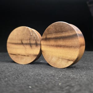 May include: Two wooden ear plugs with a natural wood grain pattern. The plugs are circular and have a light brown colour with darker brown streaks. The plugs are displayed on a dark surface.