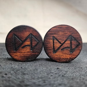 以下が含まれることがあります： 彫刻された、様式化された矢印のデザインが施された、2つの丸い濃い茶色の木製プラグ。 木目は見え、プラグはわずかに面取りされたエッジを持っています。 背景は暗く、質感のある表面です。
