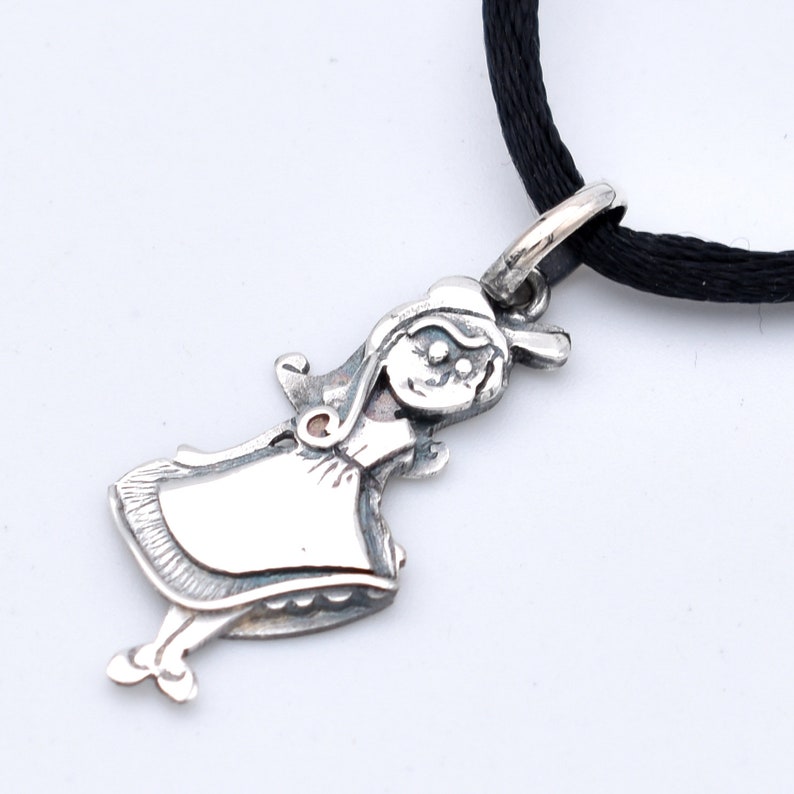 May include: A silver pendant in the shape of a cartoon girl, with a smiling face and a dress. The pendant is attached to a black cord. The girl pendant is a charming accessory.