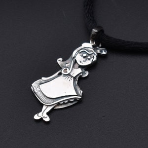 May include: A silver pendant necklace featuring a stylized girl in a dress. The pendant is attached to a black cord. The girl has a smiling face and is wearing a dress with a blank rectangular space on the front.