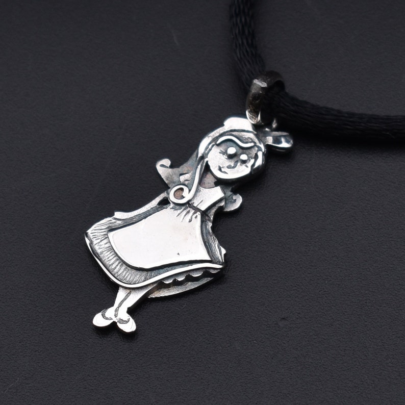 May include: A silver pendant necklace featuring a stylized girl in a dress. The pendant is attached to a black cord. The girl has a smiling face and is wearing a dress with a blank rectangular space on the front.