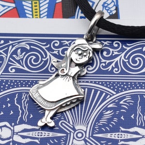 May include: A silver pendant in the shape of a cartoon girl wearing a dress and bonnet, suspended from a black cord. The pendant has detailed features and is set against a blue and white patterned background.