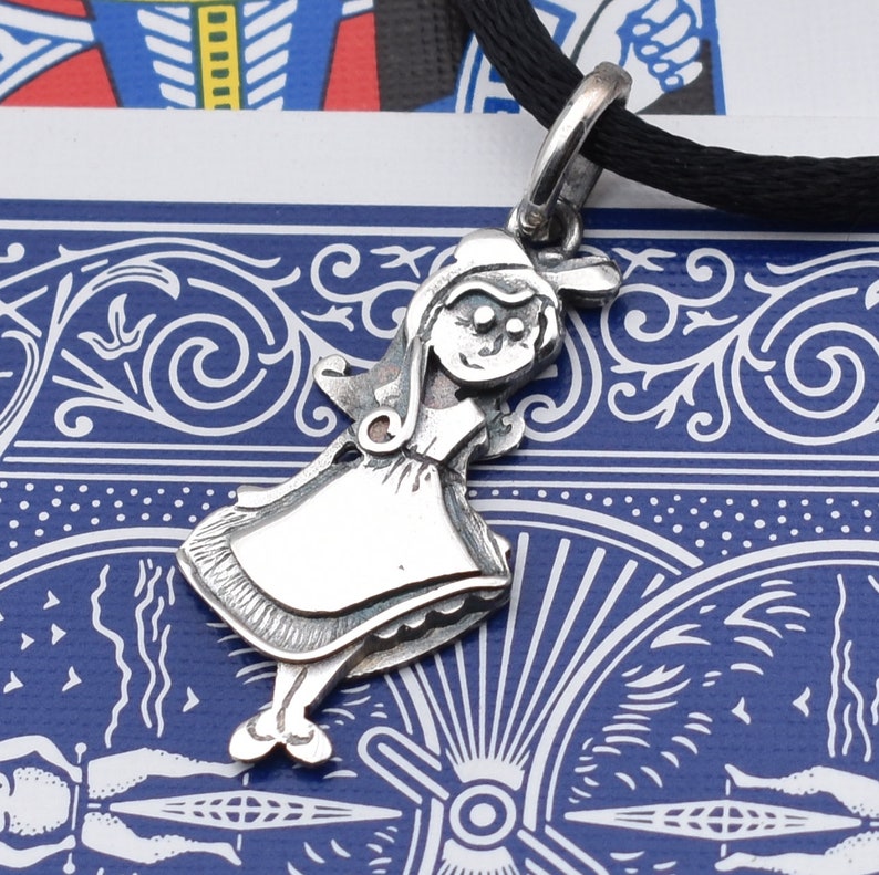 May include: A silver pendant in the shape of a cartoon girl wearing a dress and bonnet, suspended from a black cord. The pendant has detailed features and is set against a blue and white patterned background.