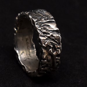 Op de afbeelding: Een getextureerde zilveren ring met een ruw, organisch ontwerp. Het oppervlak van de ring heeft een ruwe, ongelijke textuur, waardoor een unieke, handgemaakte uitstraling ontstaat. De ring staat tegen een donkere achtergrond, wat de metaalglans en ingewikkelde details benadrukt.