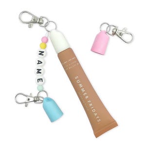May include: A cosmetic product tube with the text "SUMMER FRIDAYS" in a light brown color. The tube is attached to a keychain with a beaded strand spelling "NAME". Additional keychains with pink and blue caps are also present.