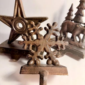 Op de afbeelding: Close-up van drie gietijzeren kerstsokkenhouders. De houders hebben de vorm van een ster, een sneeuwvlok en een scène met bomen en beren. De houders zijn allemaal donkerbruin.