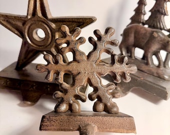 May include: Close-up of three cast iron Christmas stocking holders. The holders are in the shape of a star, a snowflake, and a scene with trees and bears. The holders are all a dark brown color.