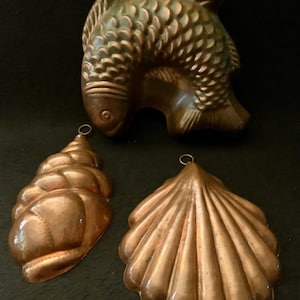 May include: Three vintage copper cake molds in the shape of a fish, a seashell, and a conch shell. The molds are all in good condition and have a beautiful patina.