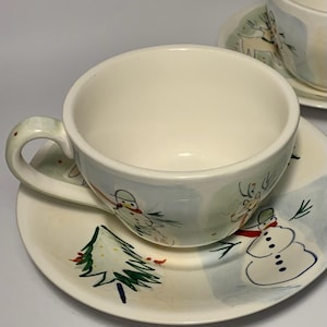 May include: Cream-colored teacup and saucer set with a winter holiday theme. The cup and saucer feature hand-painted snowmen, a Christmas tree, and a light blue and white background. The cup has a curved handle.