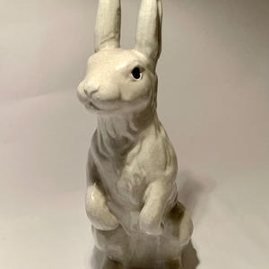 May include: A ceramic rabbit figurine in a standing pose. The off-white bunny has long ears, a detailed face, and is standing on its hind legs. The figurine has a crackle glaze finish.