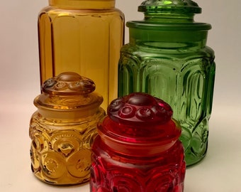 May include: Four glass jars with lids in different colors. The jars are amber, green, and red. The jars have a textured, starburst pattern.