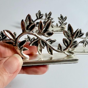 May include: Silver napkin rings featuring a detailed leaf design. Each ring has a rectangular base with a three-dimensional leaf and vine pattern. The leaves are intricately shaped, adding a touch of elegance to any table setting.