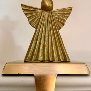 Könnte beinhalten: Ein goldfarbener Strumpfhalter aus Metall mit einer stilisierten Engelsfigur, die ein strukturiertes, plissiertes Gewand und Flügel aufweist. Die Basis besteht aus einer flachen, rechteckigen Platte mit glatter Oberfläche und einem Haken.