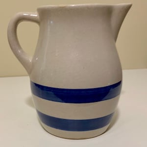 Puede incluir: Una jarra de cerámica blanca con un diseño de rayas azules y blancas. La jarra tiene un cuerpo redondeado y un asa.