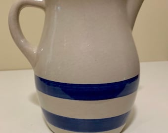 Puede incluir: Una jarra de cerámica blanca con un diseño de rayas azules y blancas. La jarra tiene un cuerpo redondeado y un asa.