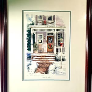 Puede incluir: Una impresión en acuarela enmarcada de una casa decorada para Navidad. La casa tiene un porche con un árbol de Navidad y una corona en la puerta. La impresión está enmarcada en un marco de madera marrón oscuro. El texto "Deck the Halls" está en la parte inferior.
