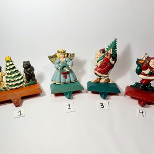 May include: Four metal Christmas stocking holders numbered one to four. The first features a deer, tree, and bear on an orange base. The second is an angel with a wreath on a green base. The third is Santa with a tree on a green base. The fourth is Santa with a sack on a red base.