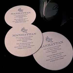 May include: Three round brown paper coasters with the instructions for making a Manhattan cocktail. Each coaster has a different design with a glass and bottle illustration. The text on the coasters reads "MANHATTAN" and includes the ingredients and instructions for making the cocktail.