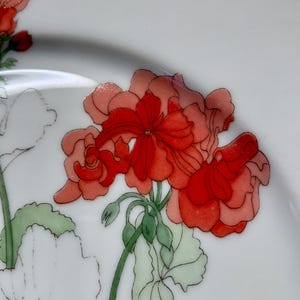 Puede incluir: Un plato de cerámica blanco con un diseño floral con geranios rojos y rosas con hojas verdes.