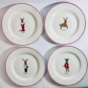 May include: Set of four white ceramic plates with a red rim. Each plate features a cartoon reindeer character with a name underneath: Vixen, Prancer, Dasher, and Dancer. A festive holiday theme.