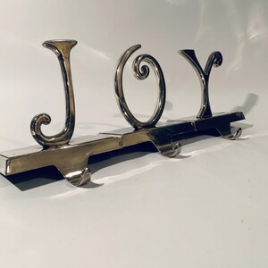 Puede incluir: Porta calcetines de metal plateado con la palabra "JOY" en letras estilizadas. Cada letra tiene un gancho para colgar calcetines.