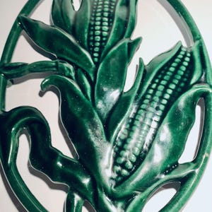 May include: Green ceramic corn on the cob hot pad. The hot pad is shaped like two ears of corn with leaves. It has a cutout design.