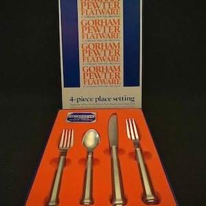 Puede incluir: Un juego de cubiertos Gorham Pewter Flatware de 4 piezas en una caja azul y roja. El juego incluye una cucharita, un cuchillo de mesa, un tenedor de mesa y un tenedor de ensalada individual. Los cubiertos están hechos de peltre y tienen un acabado pulido.