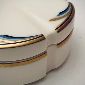 Può includere: Un portagioie decorativo bianco con un bordo smerlato. La scatola è adornata con accenti dorati e blu. Il coperchio e la base sono delineati con finiture dorate. La scatola ha una forma curva e arrotondata.