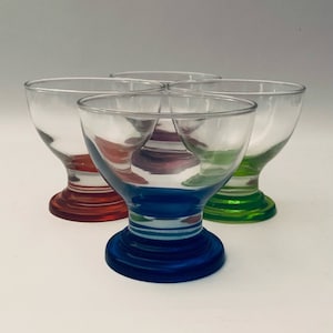Puede incluir: Juego de cuatro vasos de beber de vidrio transparente con bases de colores. Los vasos tienen un cuenco ancho y un tallo corto. Las bases son rojas, azules, verdes y moradas.