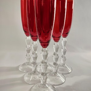 Pode incluir: Conjunto de seis taças de champanhe vermelhas. Os copos têm uma haste e uma base transparentes e facetadas. O vidro vermelho é de uma cor profunda e rica. As taças estão dispostas em semicírculo.