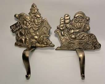 May include: Two metal Christmas stocking holders. One features a Santa Claus figure with a sleigh, and the other depicts a snowman with a Christmas tree and a birdhouse. Both have curved hooks for hanging stockings. The holders are silver in color.