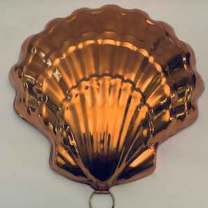 May include: A shiny copper-colored baking mold shaped like a large scallop shell. The mold features a series of radiating ridges and grooves that mimic the natural texture of a shell, with a small circular metal ring attached at the bottom edge.