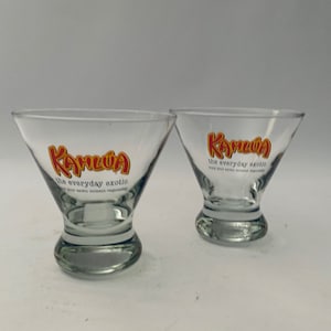 May include: Two clear martini glasses featuring the Kahlua logo in red and yellow. The text "the everyday exotic" is printed below the logo. The glasses have a flared bowl and a short, rounded stem.
