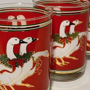 Könnte beinhalten: Set mit vier transparenten Trinkgläsern mit festlichem Design. Jedes Glas hat einen roten Hintergrund mit einer weißen Gänseillustration, verziert mit einem grünen Kranz und einer roten Schleife. Goldakzente und -verzierungen runden das festliche Thema ab.