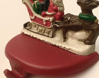 May include: A red and white ceramic Christmas stocking holder featuring Santa Claus riding in a sleigh pulled by a reindeer. The reindeer is brown with a green wreath around its neck. The sleigh is filled with presents and Santa is wearing a red suit with a white beard and hat.